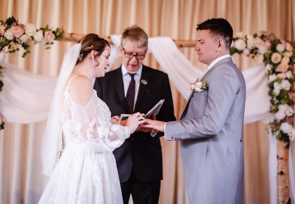 A bride and groom stand facing each other, exchanging rings during their wedding ceremony, with an officiant reading from a book behind them. The scene is decorated with flowers and white drapery.
