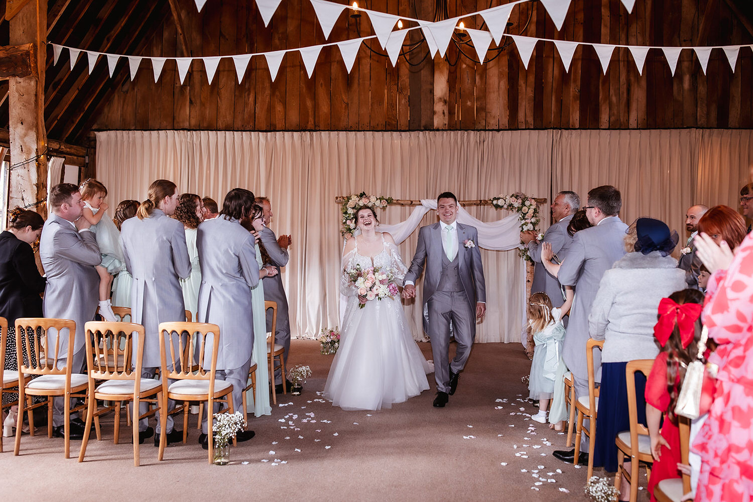 A newly married couple walks down the aisle, smiling, in a rustic venue decorated with white bunting and floral arrangements, surrounded by guests who are standing and watching.