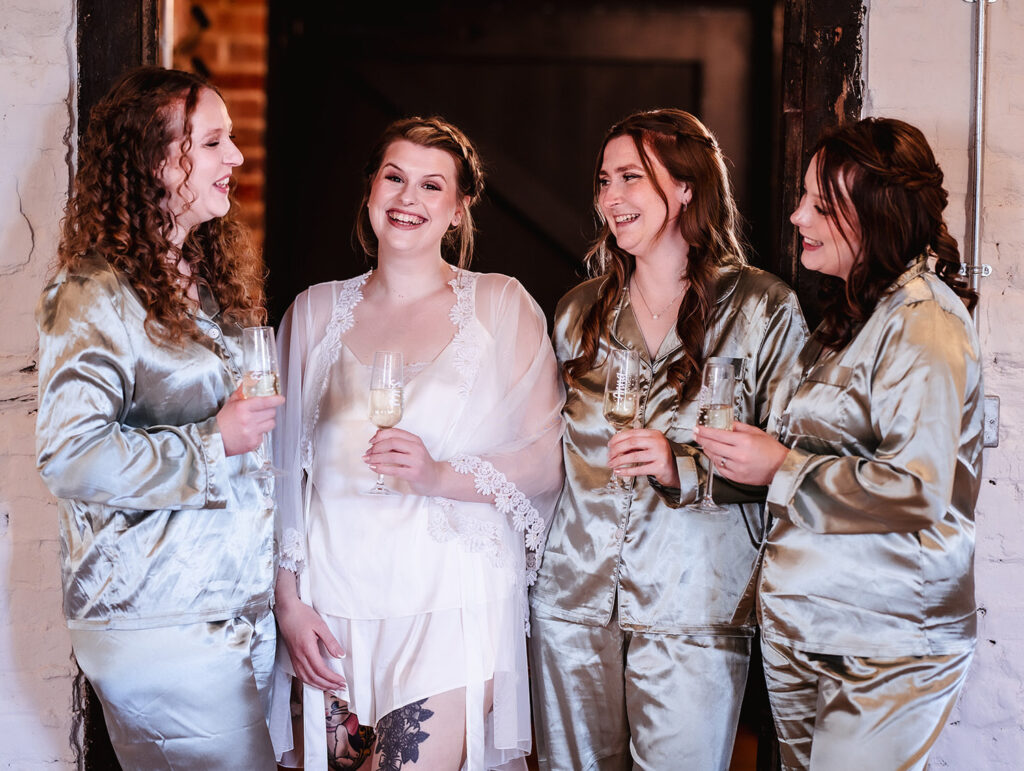 Four women stand together smiling, wearing light-colored satin pajamas, with one woman in a white robe and nightwear holding a champagne glass. They appear to be celebrating, likely before a wedding.