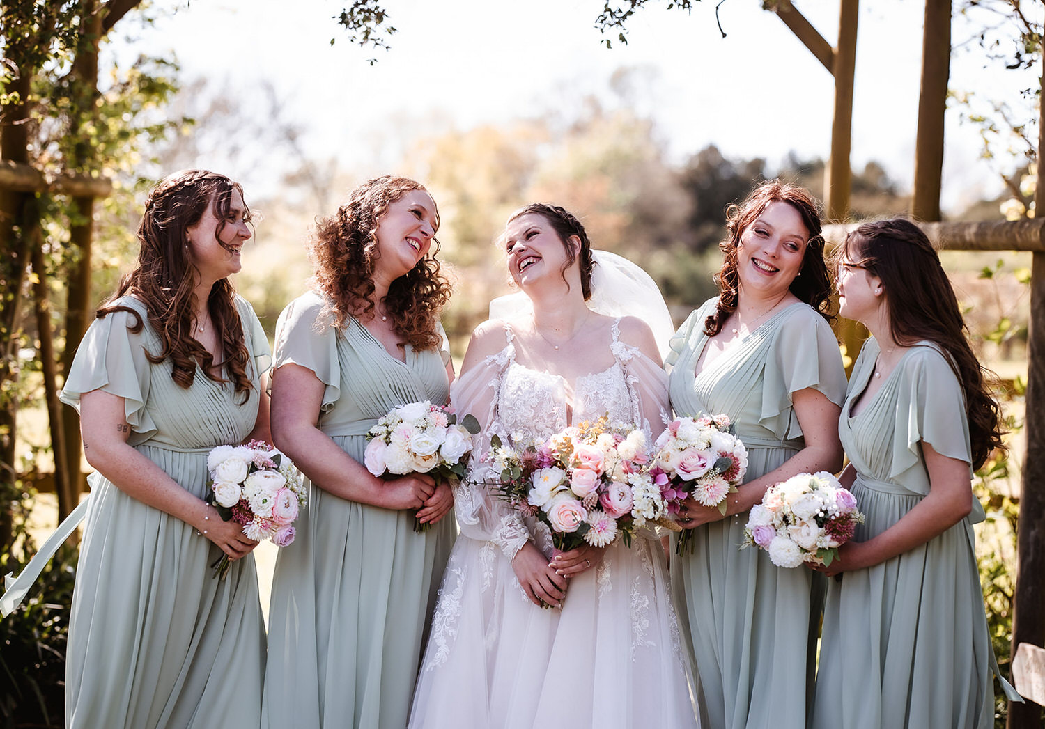 A bride in a white dress and veil smiles joyfully as she stands outdoors with four bridesmaids in matching light green dresses, all holding pastel floral bouquets. Sunlight filters through trees in the background.