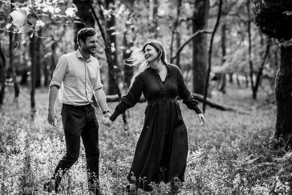 A man and woman hold hands and smile at each other while walking through a sunlit forest. The image is in black and white, capturing a joyful and romantic moment.