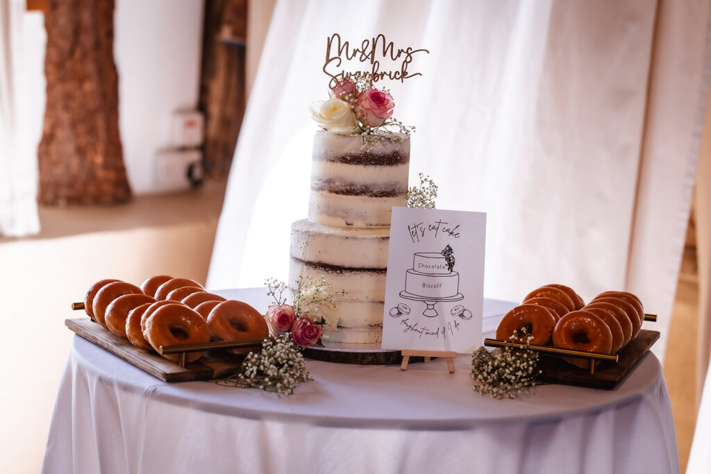 A white-frosted, semi-naked tiered wedding cake with pink and white roses sits on a round table. The cake is topped with a Mr & Mrs sign and flanked by trays of glazed donuts. A small sign describes the cake flavor.
