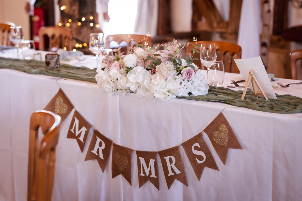 A wedding table decorated with a white cloth, floral centerpiece of white and pink flowers, and a burlap banner reading MR & MRS. Glasses, plates, and wooden chairs are arranged around the table.