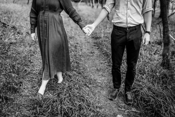 Black and white photo of a couple holding hands while walking on a grassy path. The image is cropped to show only their bodies from the shoulders down. The woman wears a long dress, and the man wears a shirt and pants.