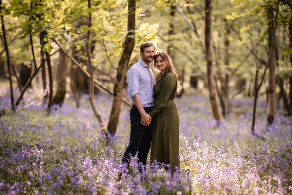 A couple stands close together, holding hands and smiling, in a sunlit woodland filled with blooming purple flowers and green-leaved trees.