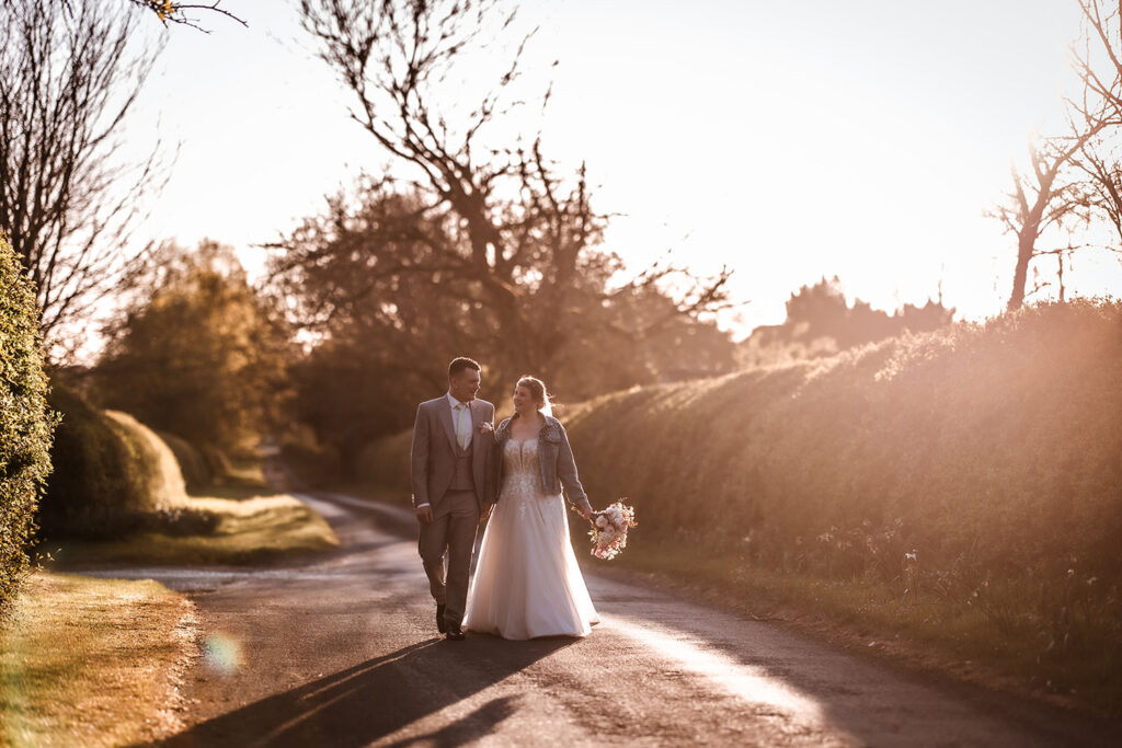A bride and groom walk hand in hand down a sunlit country lane, surrounded by hedges and bare trees, with warm, golden light creating a romantic atmosphere.