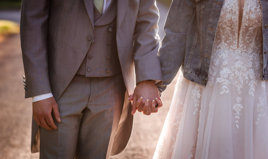 A couple holds hands outdoors; one wears a suit and the other a lace wedding dress with a denim jacket. Sunlight shines softly behind them.