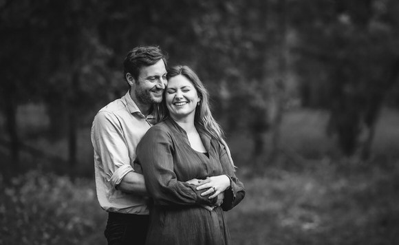 A man and woman stand close together outdoors, smiling and laughing. The man hugs the woman from behind. The image is in black and white, with a blurred natural background.
