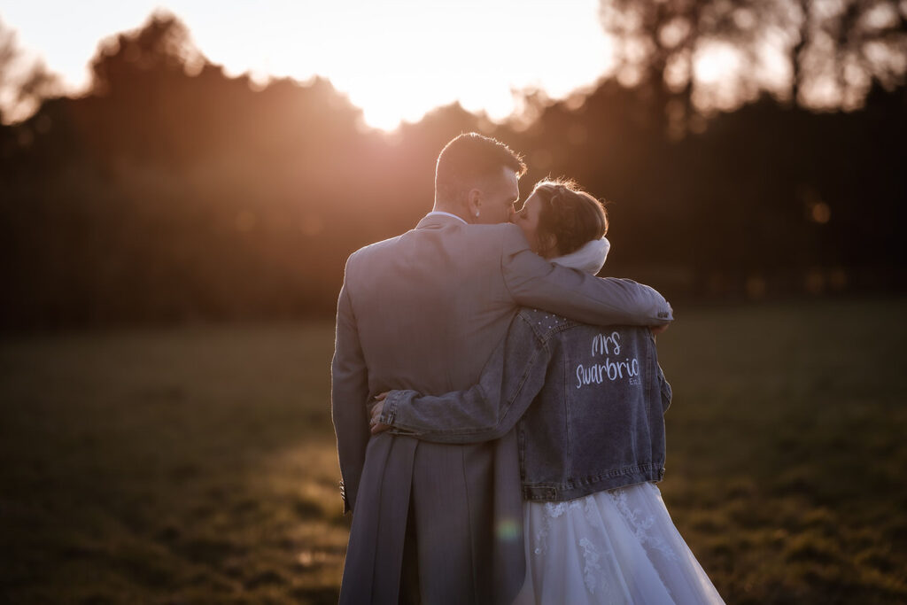 A bride and groom embrace at sunset in a field. The bride wears a denim jacket with Mrs. Sutherland on the back and a white dress, while the groom wears a gray suit. They stand close, with trees silhouetted in the background.