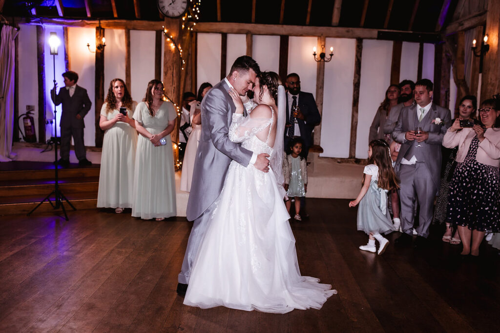 A bride and groom share their first dance at a wedding reception, surrounded by guests watching and taking photos. The room has wooden floors, rustic beams, and warm lighting.