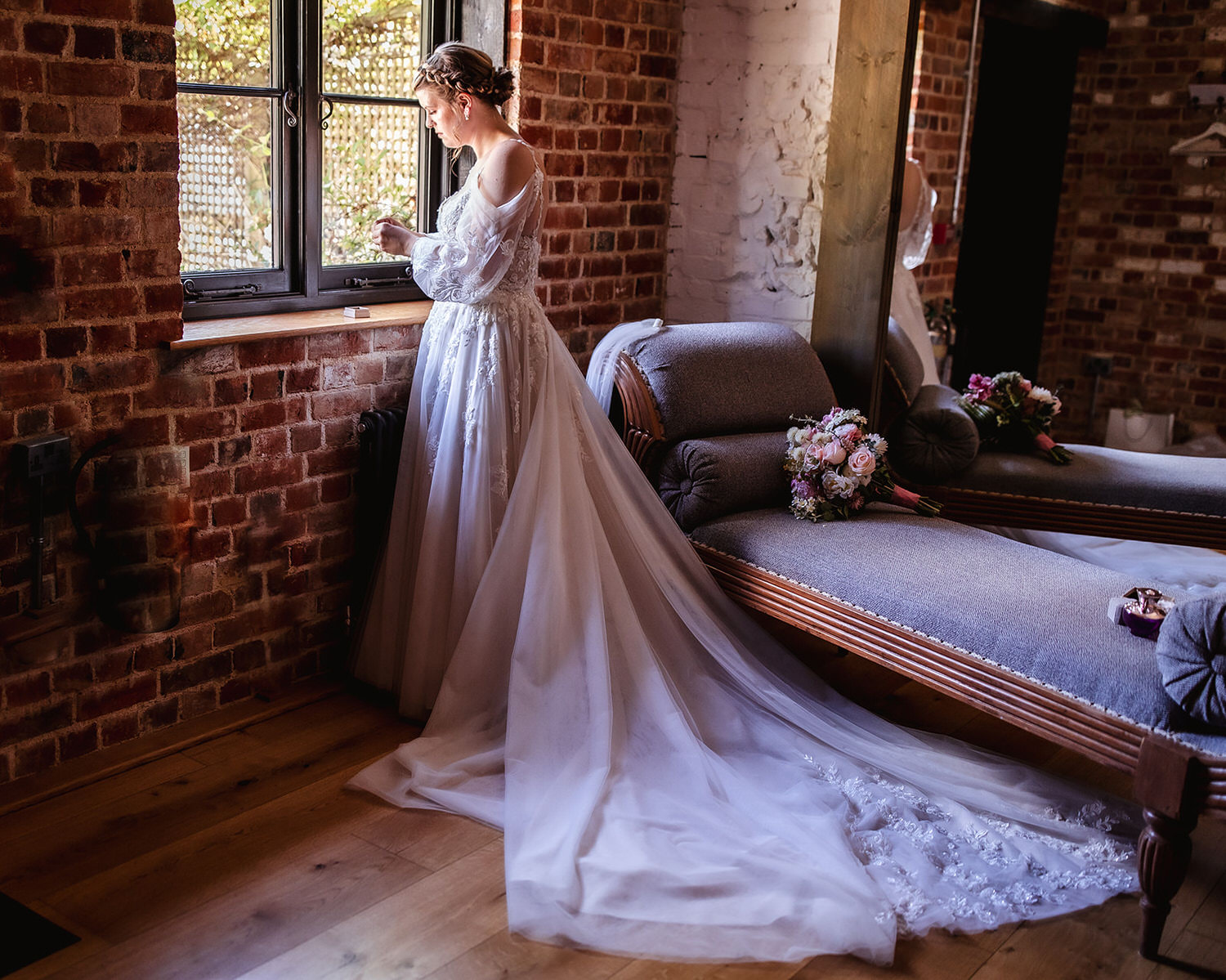 A bride in a long white gown stands by a window in a rustic room with brick walls, looking outside. Two chaise lounges and a bouquet of flowers are nearby, and sunlight streams in through the window.