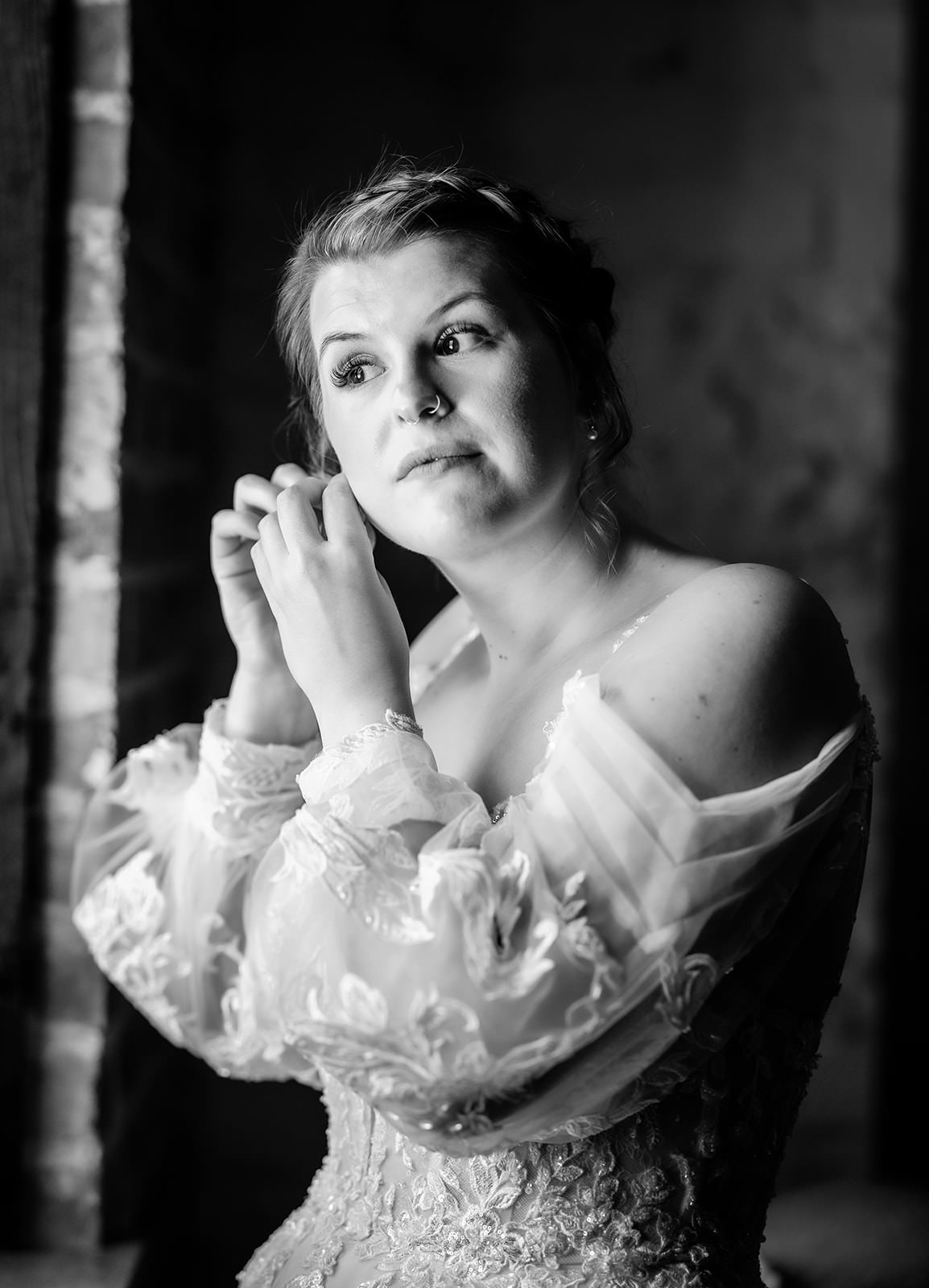 Black and white photo of a bride in a lace dress adjusting her earring while gazing thoughtfully out of a window, with soft light highlighting her face and intricate gown details.