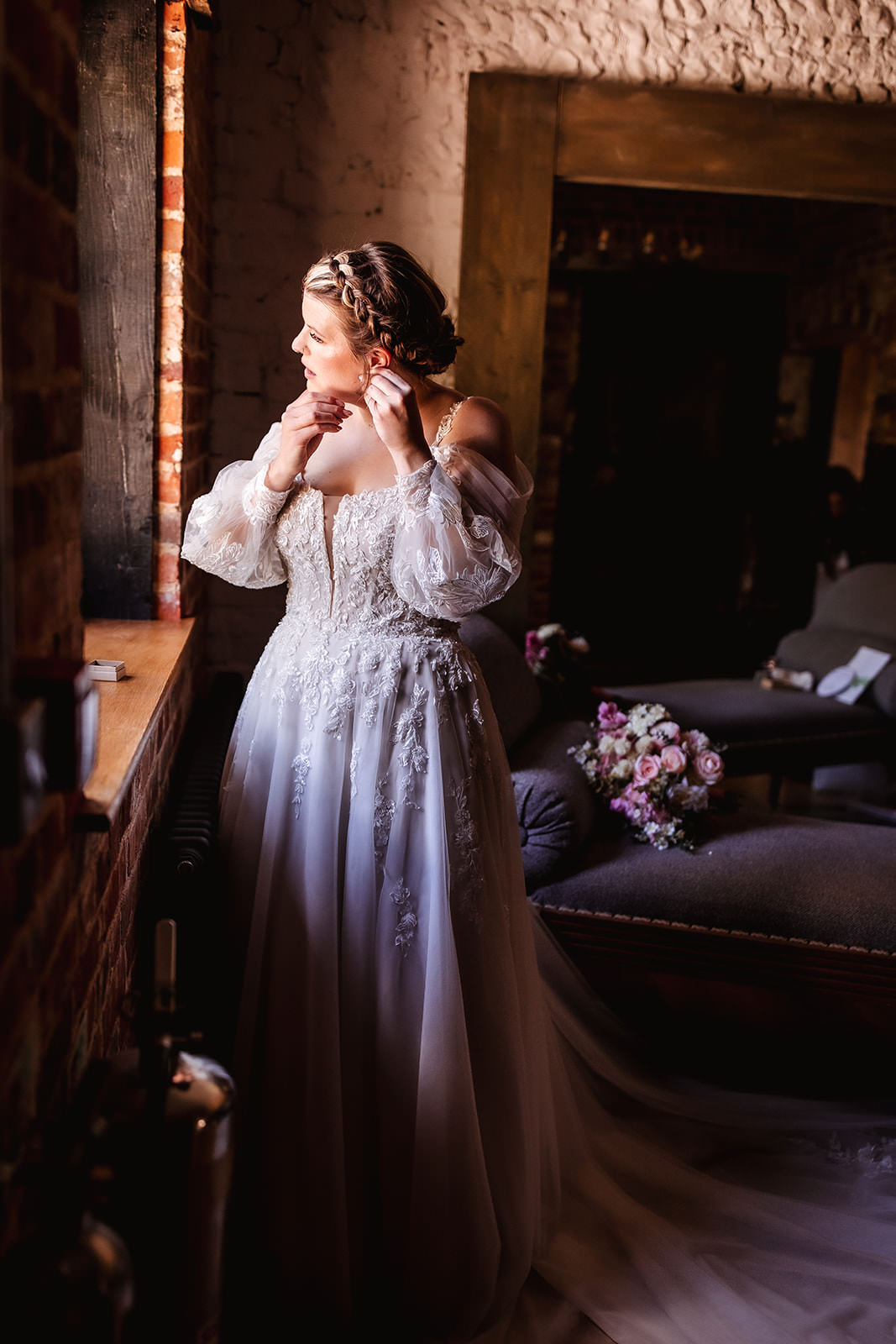 A bride in a white lace wedding dress stands by a window, adjusting her earring. Soft light shines on her as she looks outside, with a bouquet of flowers resting on a sofa in the background.