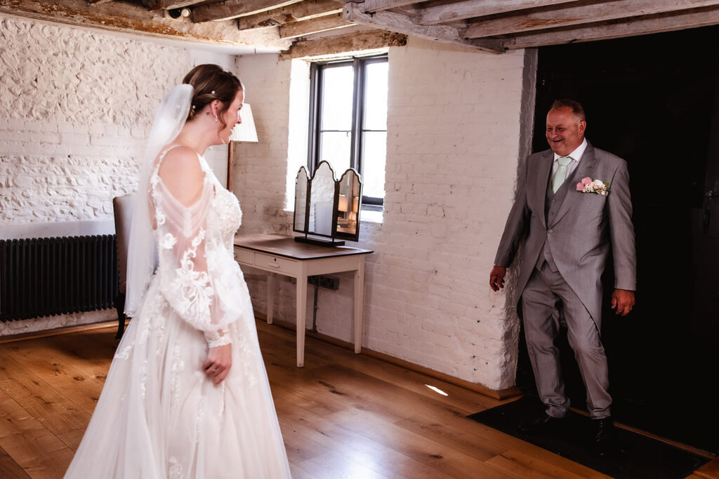 A bride in a white wedding dress and veil smiles at a man in a light gray suit and mint green tie, who looks happily surprised as he enters a rustic, sunlit room with white brick walls and wooden floors.