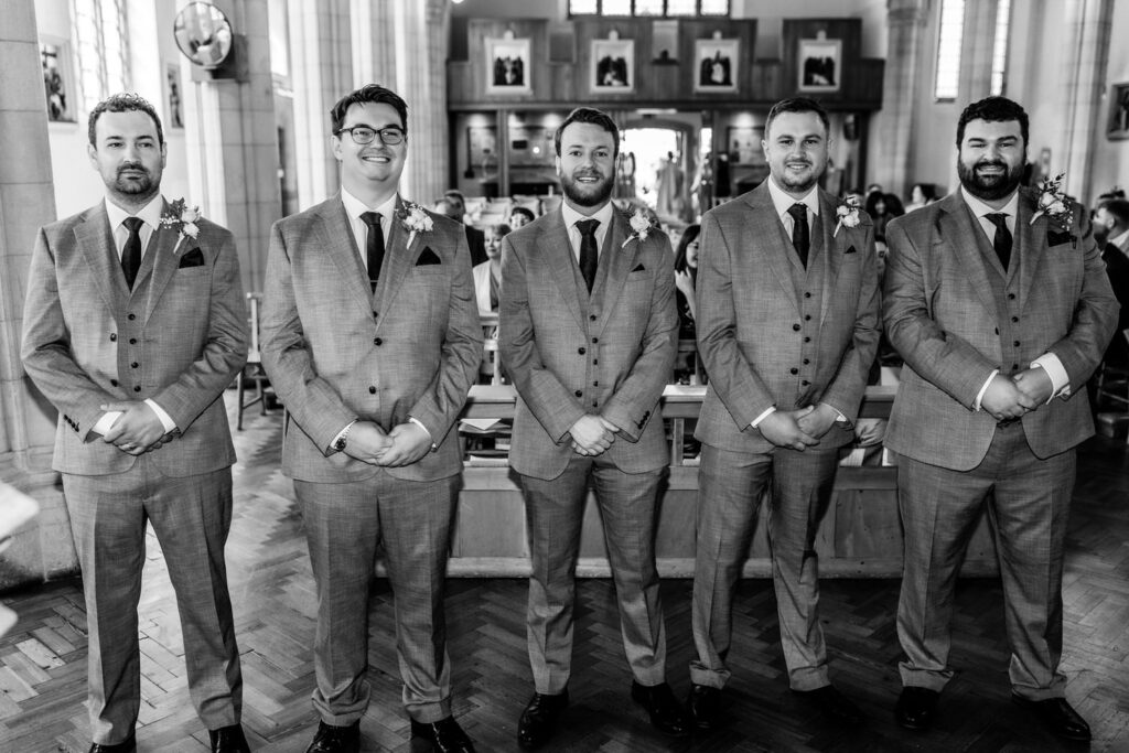 Five men in matching light-colored suits and boutonnieres stand side by side smiling inside a church, with pews and people blurred in the background.