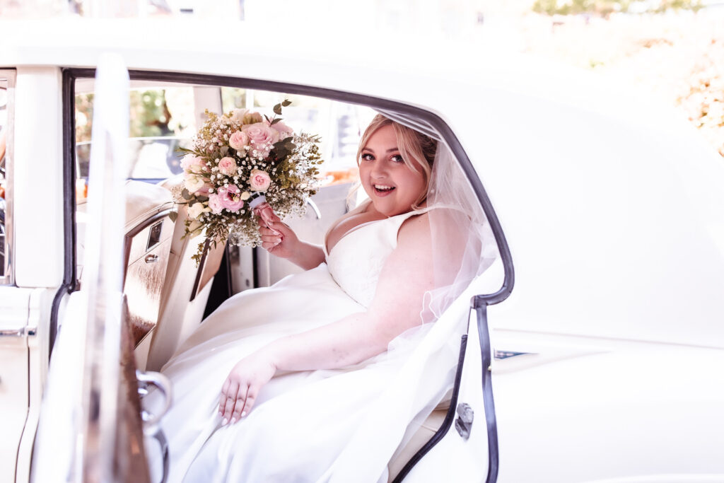 A bride in a white dress and veil sits in the backseat of a classic white car, smiling and holding a bouquet of pink and white flowers.
