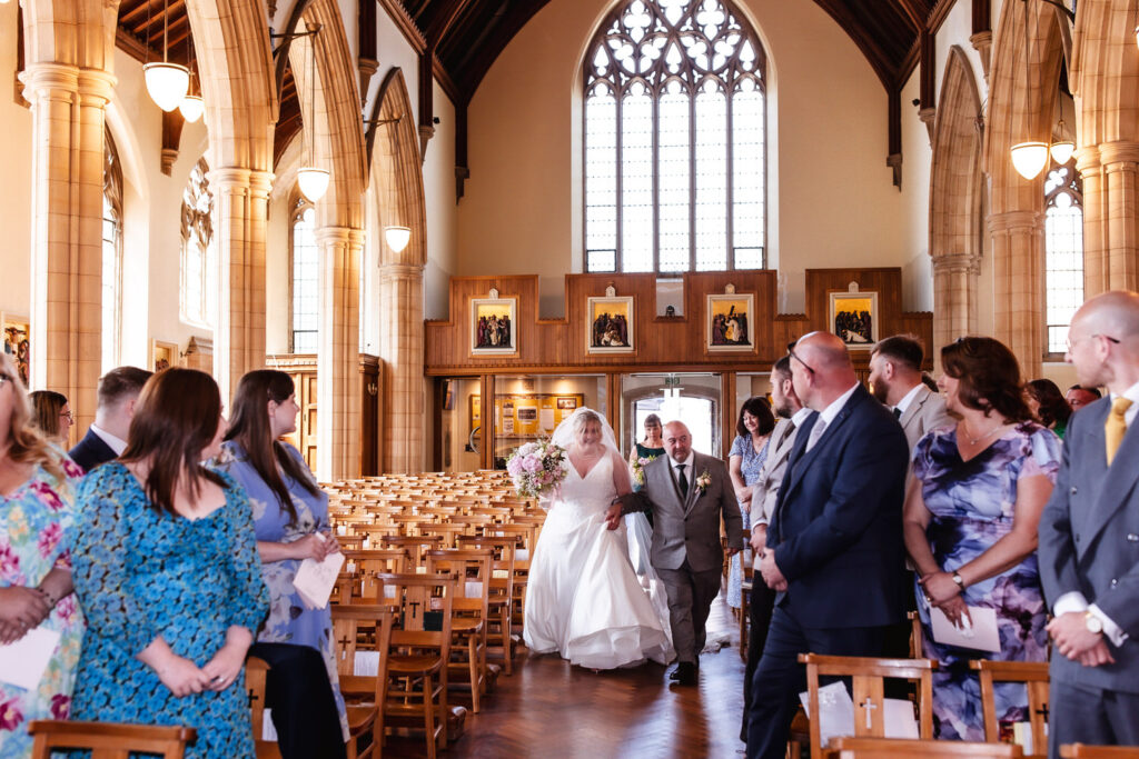 A bride in a white gown walks down the aisle of a church with high arches, accompanied by an older man. Guests in colorful attire stand and watch as they proceed toward the altar. Light streams through large windows behind them.