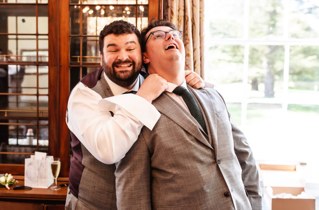 Two men in suits laugh together indoors, with one playfully grabbing the other’s necktie from behind. Sunlight streams through a large window, and a glass of champagne sits nearby. Both appear to be enjoying the moment.