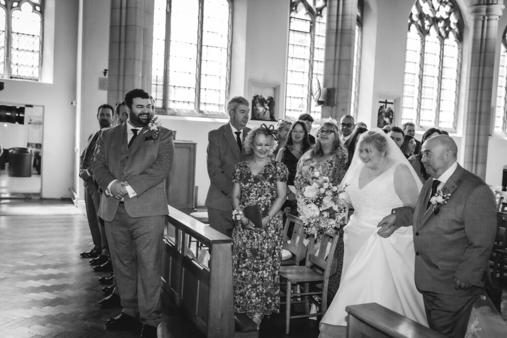 A bride in a white dress and veil walks down the aisle with an older man, smiling, surrounded by guests in formal attire inside a church with tall, arched windows.