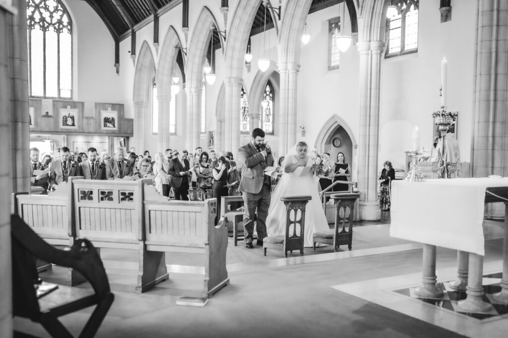 A bride and groom kneel at the altar in a church during their wedding ceremony, with guests seated and standing in the background. The scene is in black and white, highlighting the churchs elegant architecture.