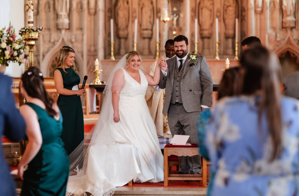 A bride and groom, smiling and holding hands, stand at the altar in a church surrounded by wedding guests. The bride wears a white gown and veil; the groom is in a gray suit. People in colorful attire look on and take photos.