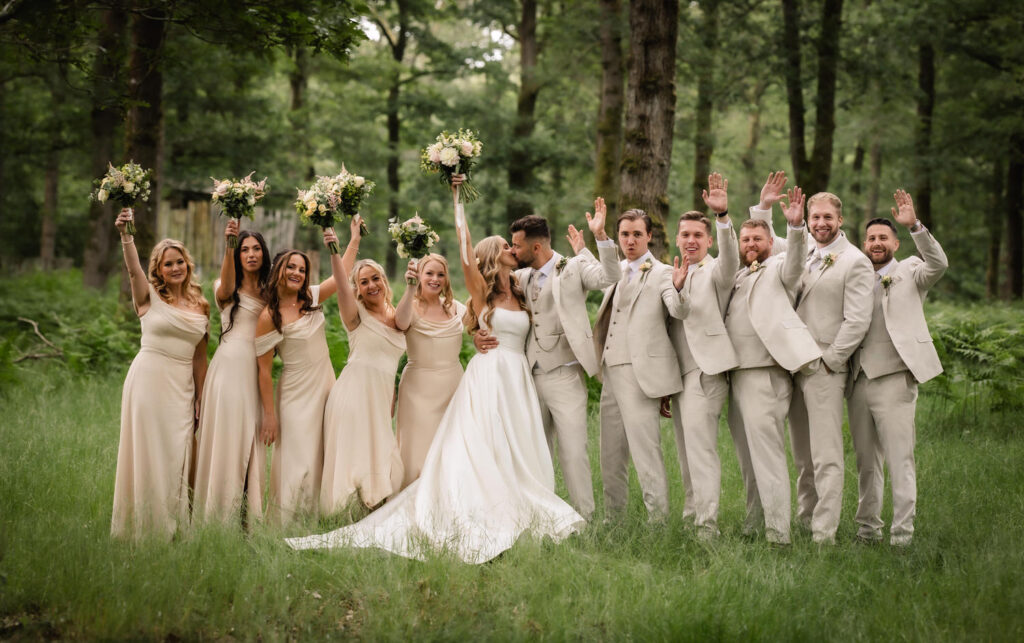 A wedding party poses outdoors in a lush green forest. Captured by a Hampshire wedding photographer, the bride and groom stand in the center, surrounded by joyful bridesmaids and groomsmen with raised bouquets and hands.