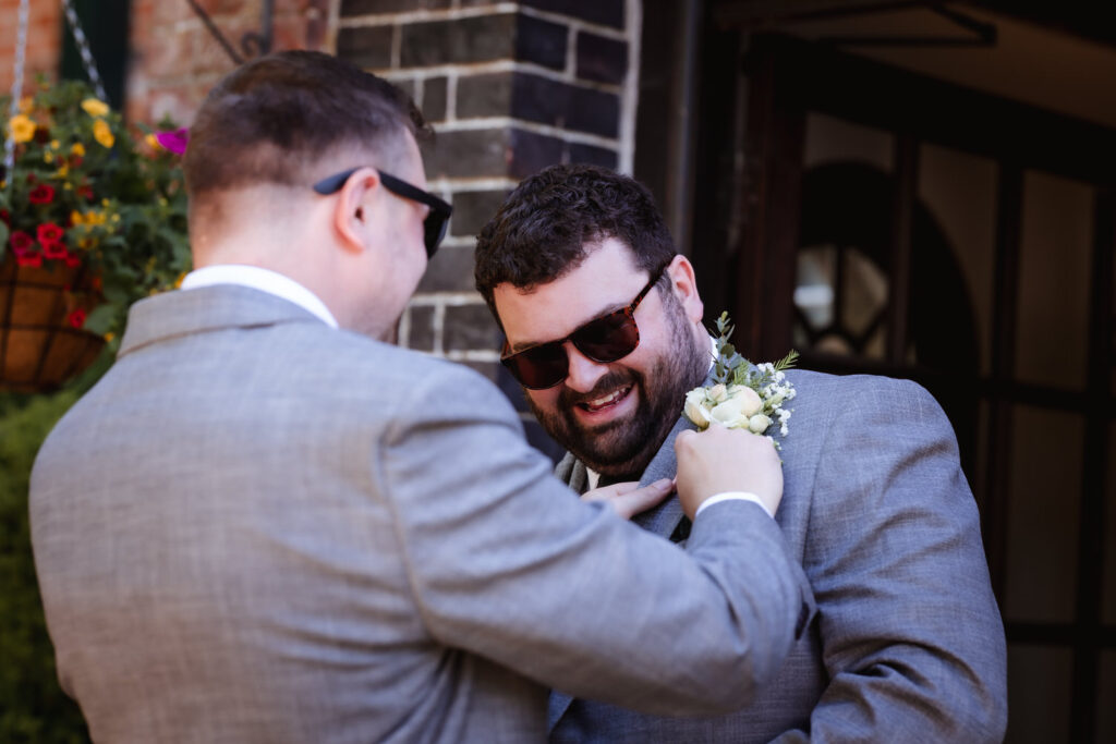 Two men in gray suits and sunglasses smile as one helps the other adjust a boutonniere on his lapel. They are standing outdoors near a brick wall and hanging flowers.