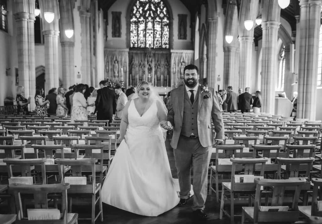 A joyful bride and groom, holding hands and smiling, walk down the aisle in a church filled with guests and rows of wooden chairs. The scene is in black and white, and the church interior is grand and ornate.