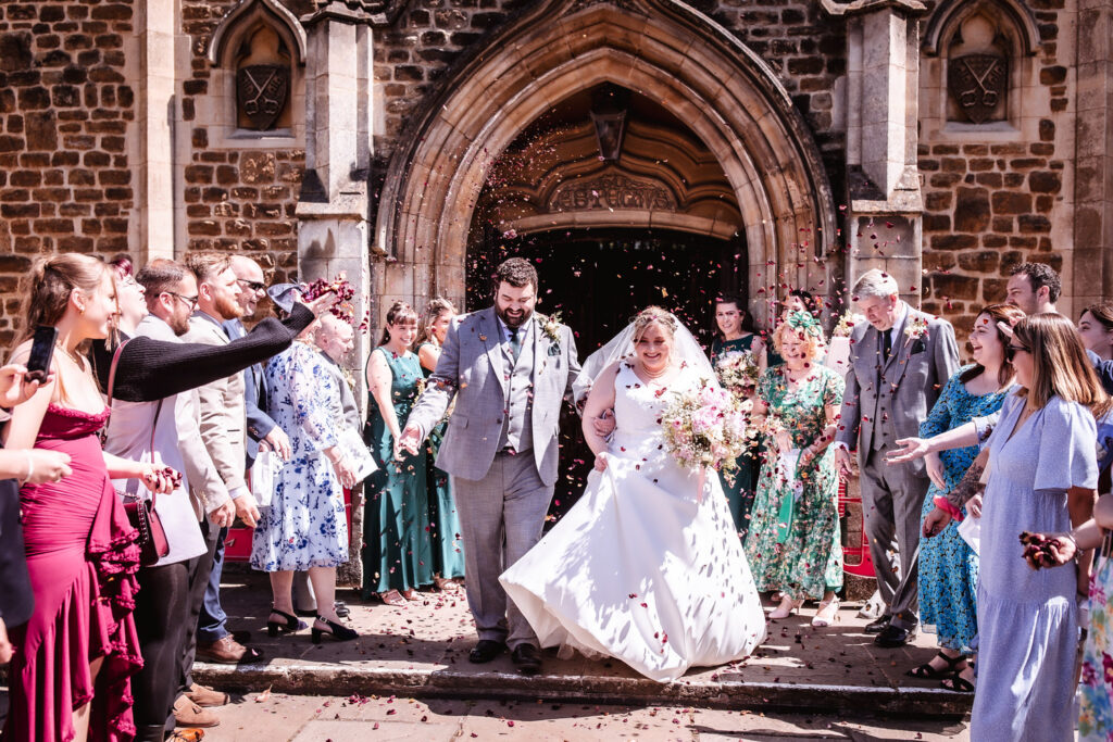 A newlywed couple exits a church, smiling as guests on either side throw flower petals. The bride wears a white dress and the groom a light gray suit. Family and friends, dressed formally, celebrate the wedding in bright sunlight.