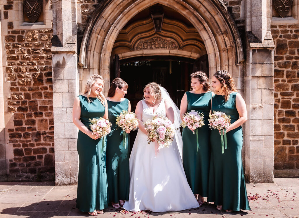 A bride in a white gown stands smiling with four bridesmaids in matching teal dresses, each holding a bouquet, in front of a stone church entrance.