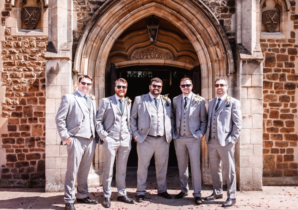 Five men in matching light gray suits and sunglasses stand smiling outside a stone building with an arched doorway, likely at a wedding or formal event.