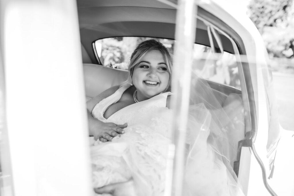 A bride in a wedding dress sits in the backseat of a car, smiling and looking out the window. The photo is in black and white, capturing a joyful, candid moment.