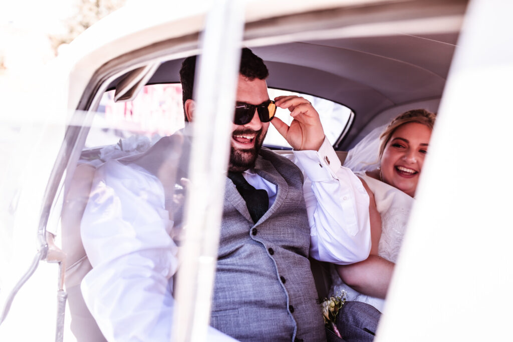 A smiling groom in a gray suit adjusts his sunglasses while sitting in the back seat of a car next to the bride, who is also smiling and dressed in white.