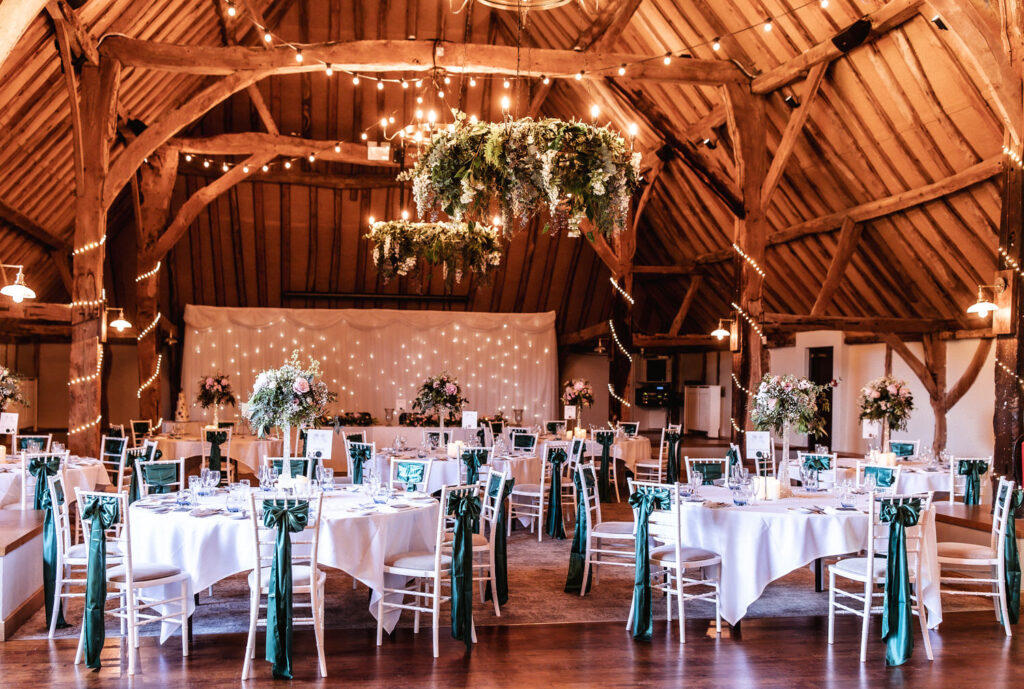 A rustic wedding reception hall with wooden beams, round tables covered in white cloths, green chair sashes, tall floral centerpieces, string lights, and hanging greenery chandeliers.
