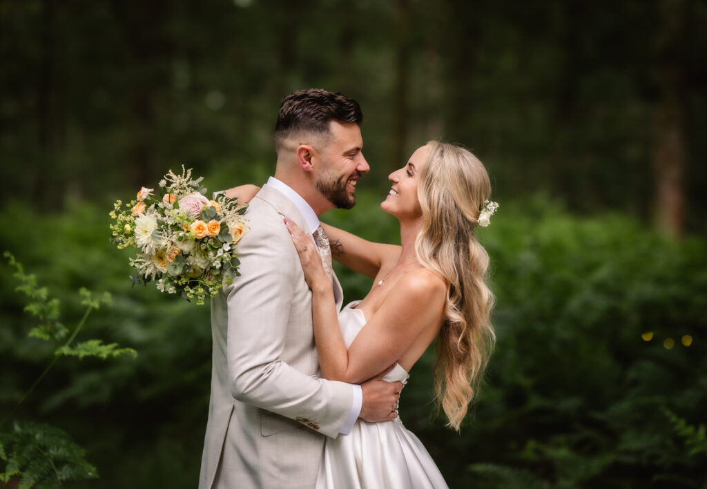 A bride and groom embrace and smile at each other outdoors, surrounded by green foliage. Captured by a Hampshire wedding photographer, the groom wears a light suit while the bride holds a bouquet of yellow and peach-toned flowers.