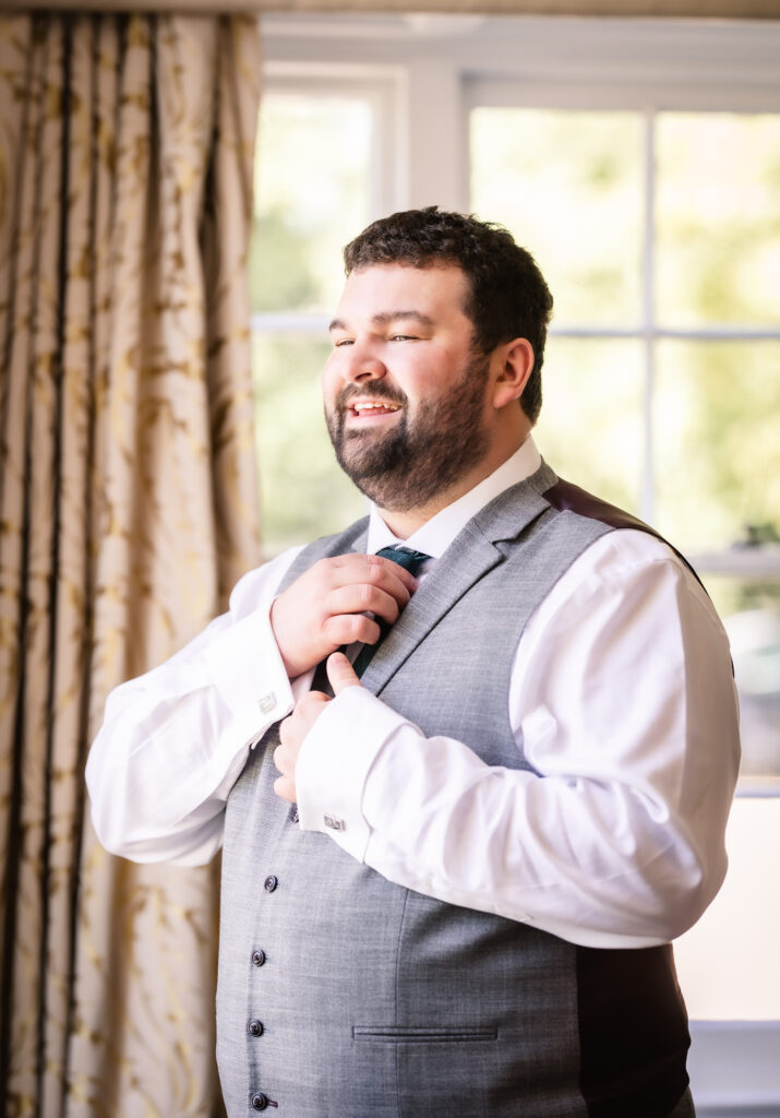 A smiling man with a beard adjusts his tie while wearing a light gray suit and white shirt, standing indoors near a sunlit window with patterned curtains.