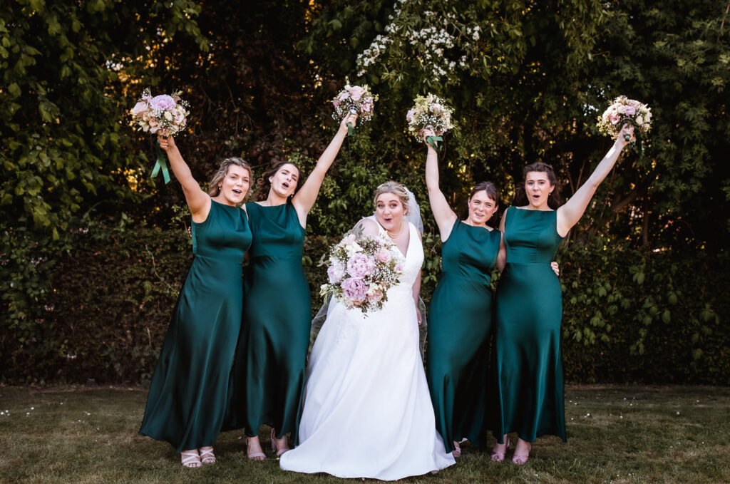 A bride in a white dress and four bridesmaids in matching green dresses stand together outdoors, smiling and raising bouquets joyfully, with lush greenery in the background.