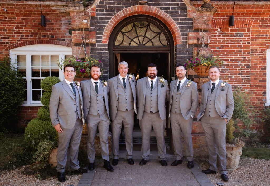 Six men in matching grey suits and ties stand smiling in front of a brick building with an arched doorway and flower baskets, likely posing for a wedding or formal event photo.