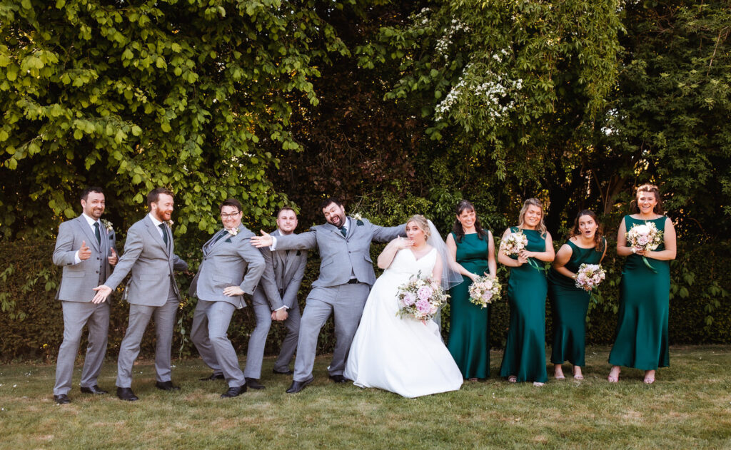A wedding party poses outside on grass in front of leafy trees. The bride in white and groom in grey stand at the center, surrounded by bridesmaids in green dresses and groomsmen in grey suits, all smiling and being playful.