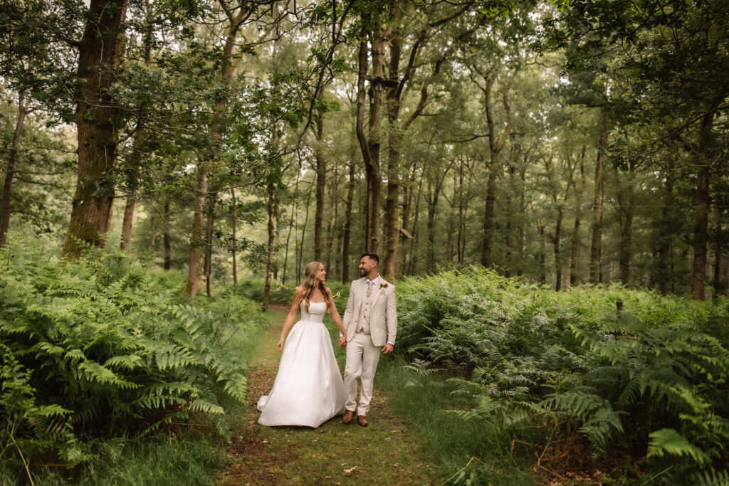A bride and groom walk hand in hand along a fern-lined path in a tranquil forest, smiling at each other. Captured by a Hampshire wedding photographer, the scene features tall trees surrounding them in a peaceful, natural setting.