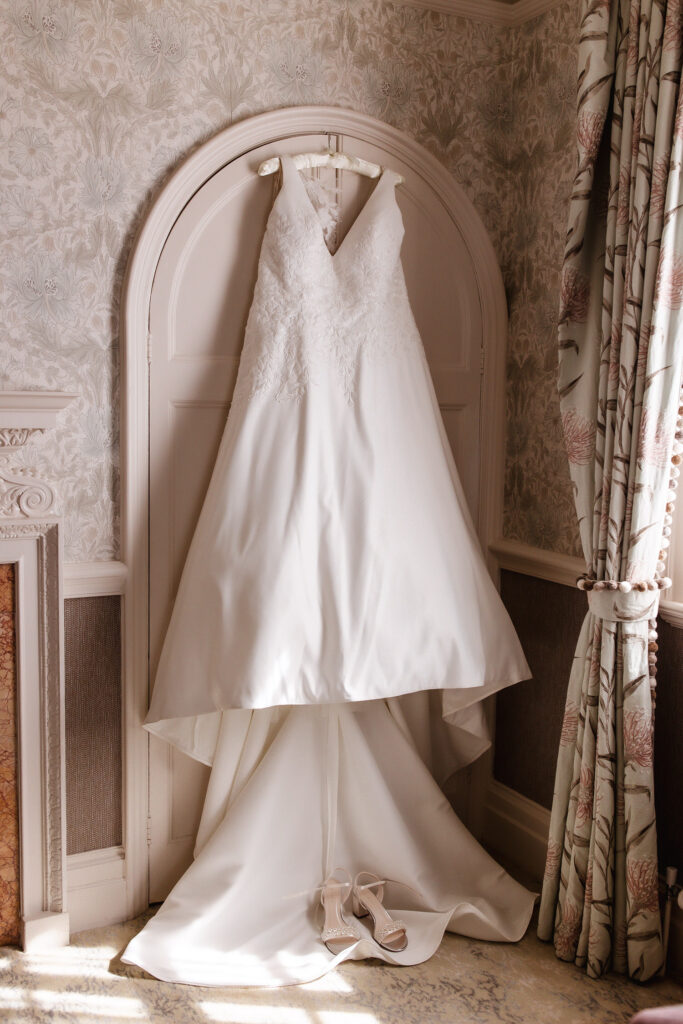 A white sleeveless wedding dress hangs on a door, with lace detailing on the bodice and a long train. A pair of matching high-heeled shoes is placed on the floor beneath the dress.