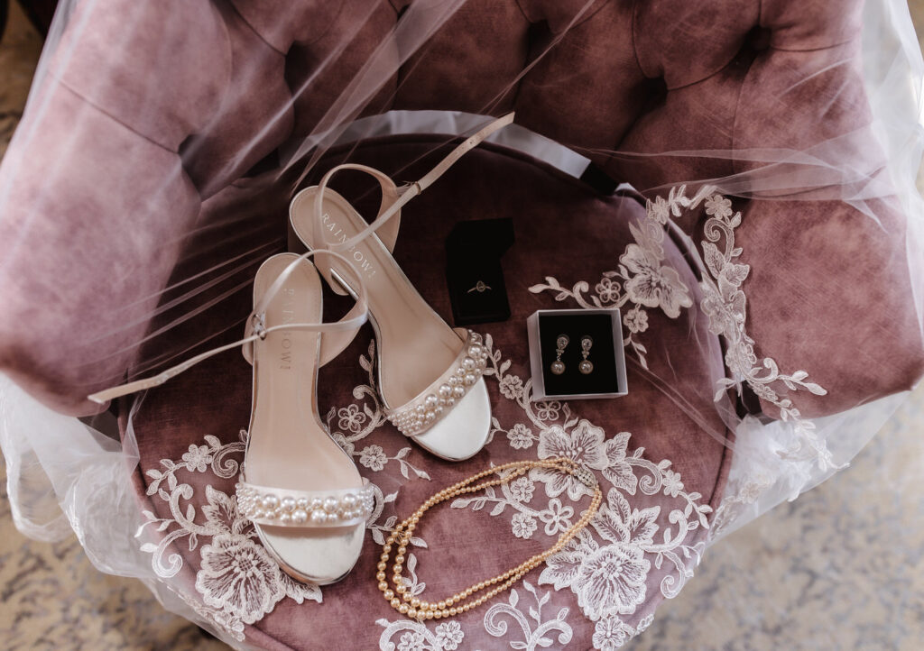 White open-toe bridal shoes, pearl necklace, earrings, and a ring box are arranged on a mauve velvet chair with a lacy white veil draped across the seat.