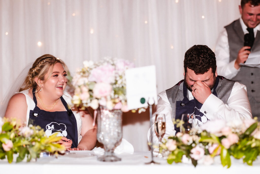 A bride and groom sit at a decorated table, both wearing navy aprons and laughing. The groom covers his face with his hand while the bride smiles. A man stands behind them with a microphone, speaking.