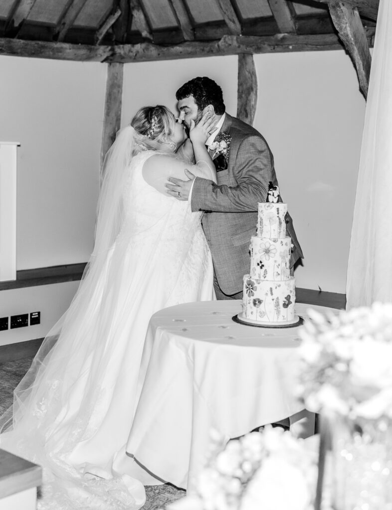 A bride and groom share a kiss beside a decorated wedding cake on a table, inside a rustic venue with exposed wooden beams. The bride wears a veil and gown; the groom is dressed in a suit.
