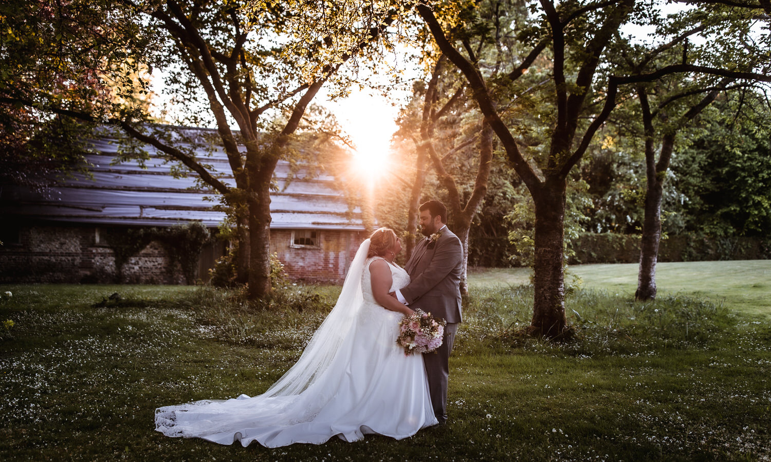 A bride and groom in wedding attire stand embracing under trees at sunset, with sunlight streaming through the branches. The bride holds a bouquet, and a rustic building is visible in the background.