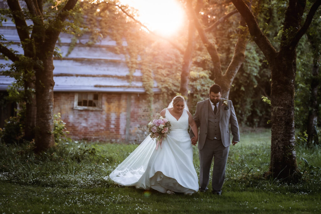 A bride and groom walk hand in hand under trees at sunset, with the bride holding a bouquet of flowers. Sunlight filters through the trees, and an old brick building stands in the background.