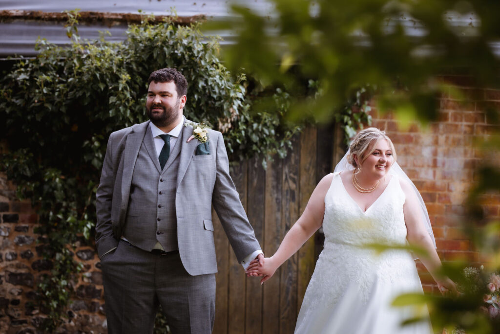 A bride and groom stand outside, holding hands and smiling. The groom wears a grey suit and dark tie; the bride wears a white dress and veil. Greenery and a rustic brick wall are in the background.