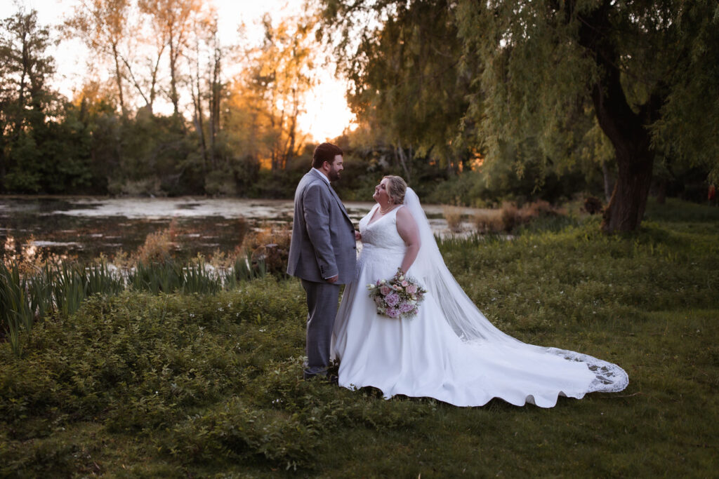 A bride and groom stand smiling at each other outdoors by a pond at sunset. The bride holds a bouquet and wears a long white gown with a veil, while the groom is in a grey suit. Trees and greenery surround them.