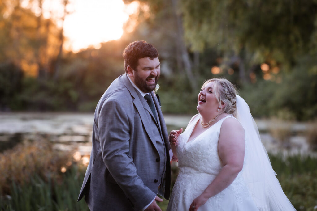 A bride and groom laugh together outdoors at sunset, standing near water with greenery in the background. The groom wears a gray suit, and the bride wears a white dress and veil. Both are smiling and joyful.