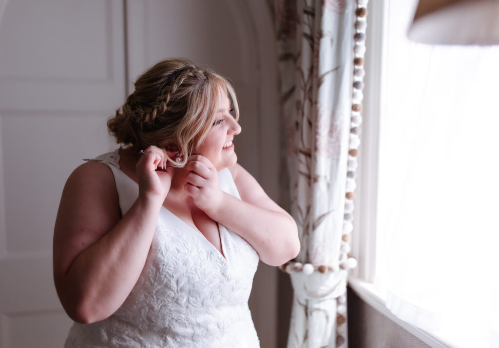 A bride in a white, sleeveless wedding dress stands by a window, smiling as she puts on an earring. Soft natural light illuminates her face and braided hair, and floral curtains frame the window.